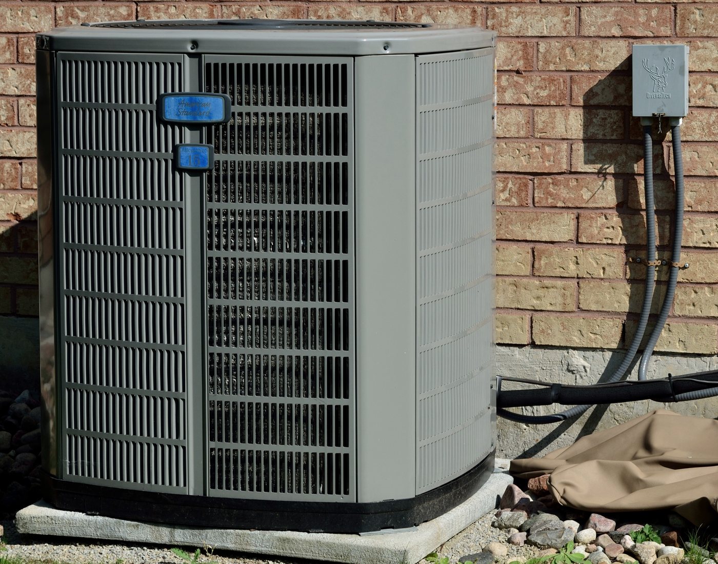 A grey residential air-conditioning condenser standing on a concrete pad against a brick house wall, with the electrical disconnect box mounted on the wall above it and the black insulated lineset running from the back of the unit into the house — the condenser plus the three add-ons a replacement quote prices separately.