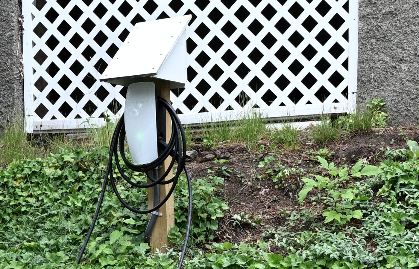 A white Tesla wall connector mounted outdoors on a wooden post in front of a lattice screen, its charging cable coiled around the post, with the unit and its mount marked — an outdoor installation where the circuit has to be run out from the house to reach the parking spot.