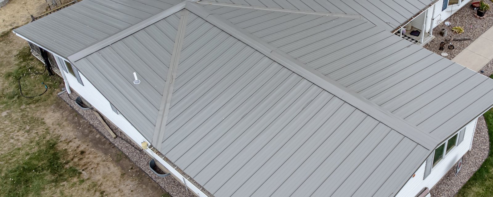 Aerial view of a single-storey house roofed in light grey metal panels, with one raised standing seam, the hip where two roof planes meet, and the eave edge along the gutter all marked — the three details a metal roofing bid is written around.