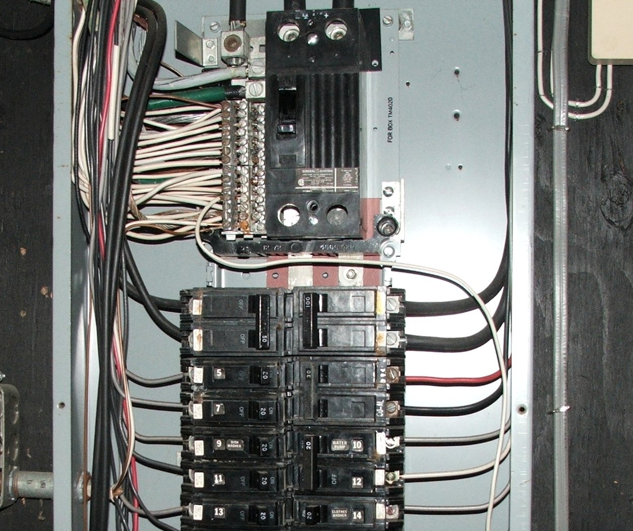 The inside of an American residential panel with its cover removed, showing the main breaker at the top of the enclosure, two columns of branch breakers filling the bus below it with a black and a white conductor leaving each one, and the grey panel box itself boxing all of it in against a dark basement wall.